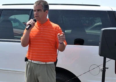 A man stands near a white SUV and speaks into a microphone during an event at Falcon Lakes Golf Course.